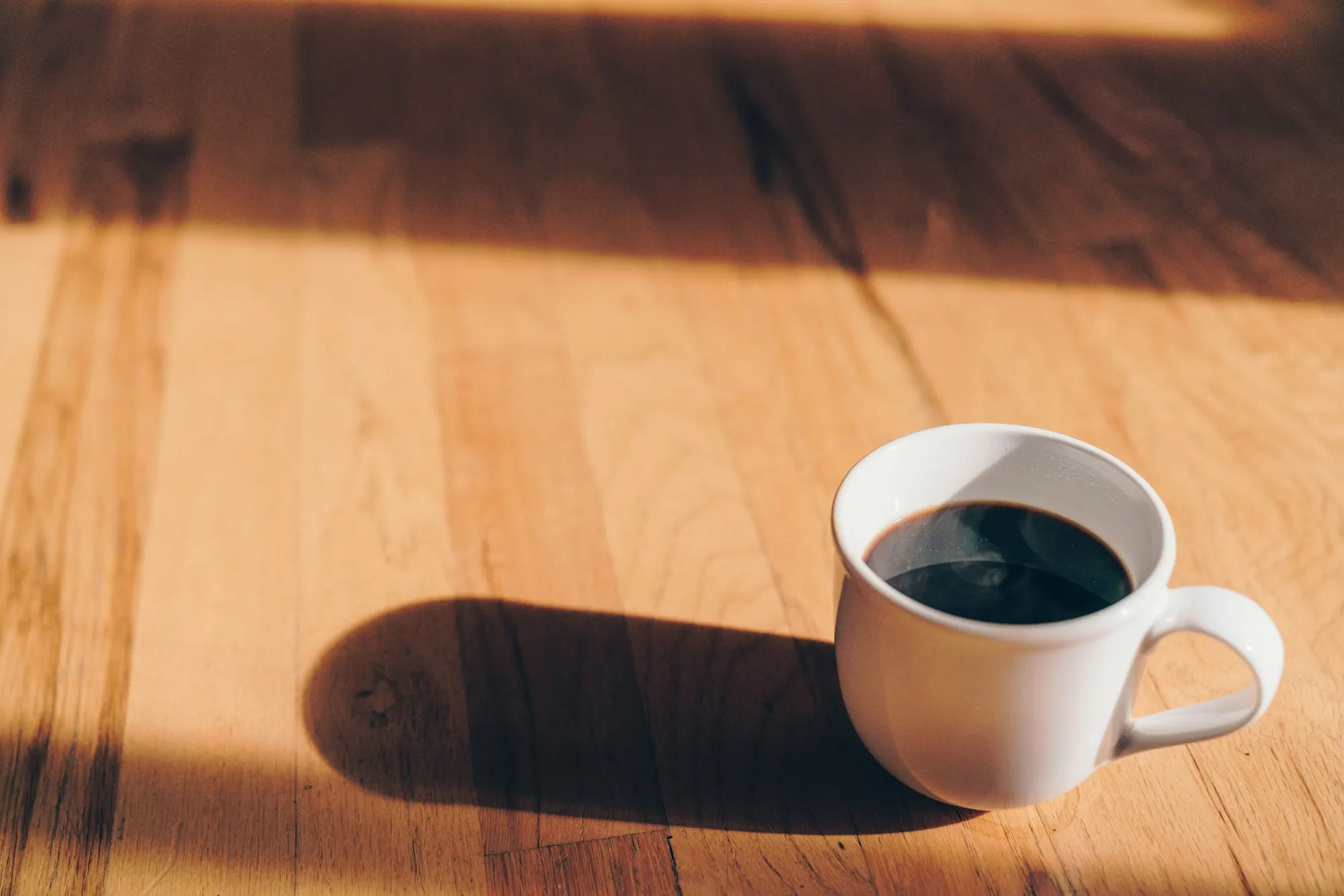 日の当たる木の床に置かれた、白いマグカップのコーヒー